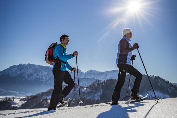 Zwei Personen mit Schneeschuhen und Wanderstöcken gehen auf verschneitem Bergweg bei Sonnenschein, im Hintergrund Bergpanorama.