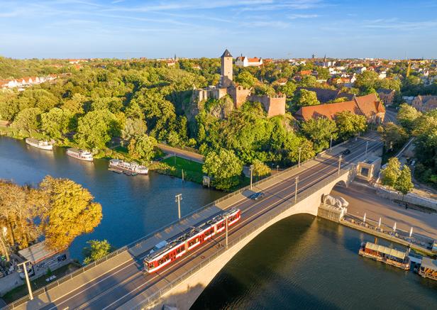 Ein Zug überquert eine Brücke, die sich über einen Fluss erstreckt, im Hintergrund befindet sich die Burg Giebichenstein.