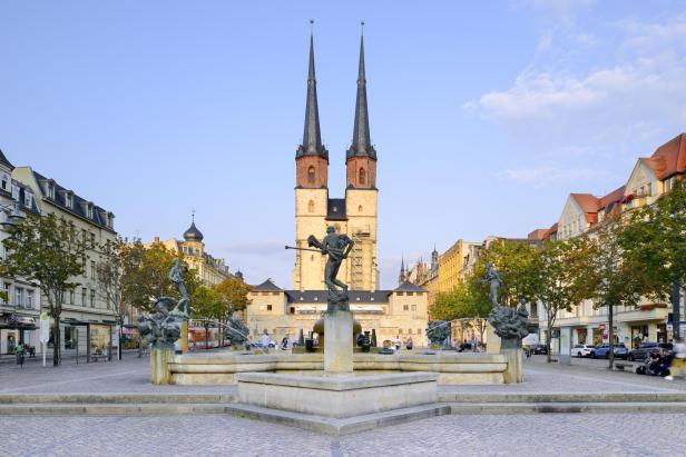Eine große Kirche mit zwei hohen Türmen steht im Hintergrund auf dem Hallmarkt in Halle (Saale). Im Vordergrund steht ein Brunnen mit der Statue einer Person, die ein Horn hält, in der Mitte, umgeben von kleineren Statuen und Wasserspielen.