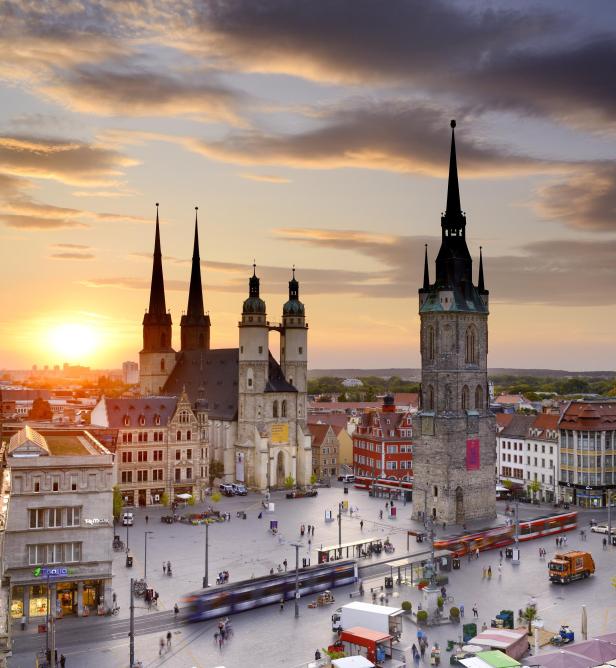 Halle (Saale), Marktplatz mit Marktkirche und Rotem Turm bei Sonnenaufgang. Der Platz ist mit Menschen, Gebäuden und einigen fahrenden Fahrzeugen bevölkert. Im Hintergrund geht die Sonne auf und erhellt den Himmel und die Wolken mit warmem Licht.