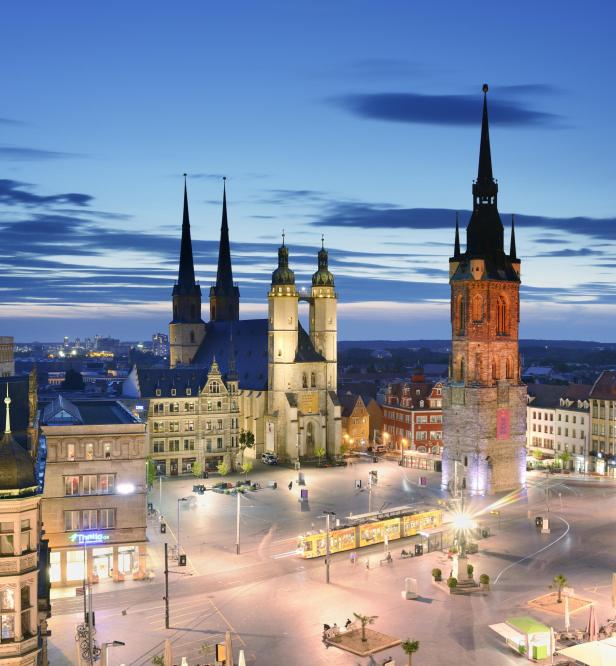 Marktplatz in Halle (Saale), bei Nacht. Die Marktkirche und der Rote Turm sind mit ihren beleuchteten Türmen zu sehen. Auf dem Platz sind Gebäude und eine Straßenbahn zu sehen. Der Himmel ist wolkenverhangen und hat einen leicht dunkelblauen Farbton. 