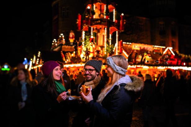 Drei Personen mit Winterkleidung essen an einem beleuchteten Weihnachtsmarktstand bei Nacht.