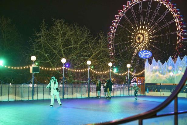 Rollschuhbahn mit mehreren Rollschuhläufern, im Hintergrund beleuchtetes Riesenrad und Lichterketten an Bäumen und Laternen.