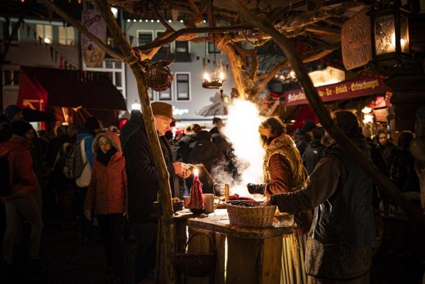 Marktstand mit brennendem Feuer und Korb, Menschen in Winterkleidung, beleuchtete Häuser im Hintergrund bei Nacht.