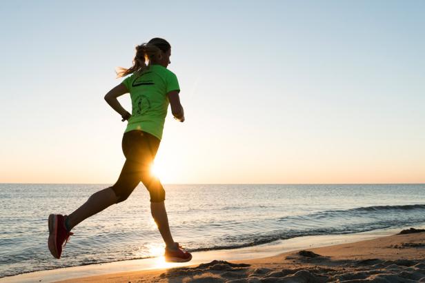 Frau in grüner Sportkleidung joggt am Strand bei Sonnenuntergang.