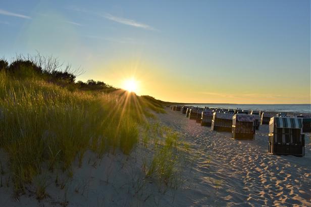 Sonnenuntergang über Sanddünen mit Strandkörben am Meer unter klarem Himmel.