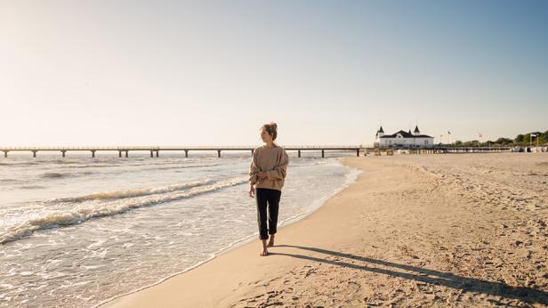 Person in beiger Jacke und dunkler Hose geht barfuß am Strand entlang, im Hintergrund eine lange Seebrücke und ein Gebäude mit Türmchen.