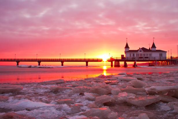 Seebrücke Ahlbeck im Winter bei Sonnenuntergang, Eisplatten auf dem Wasser, Gebäude mit Türmen am Ende der Brücke.