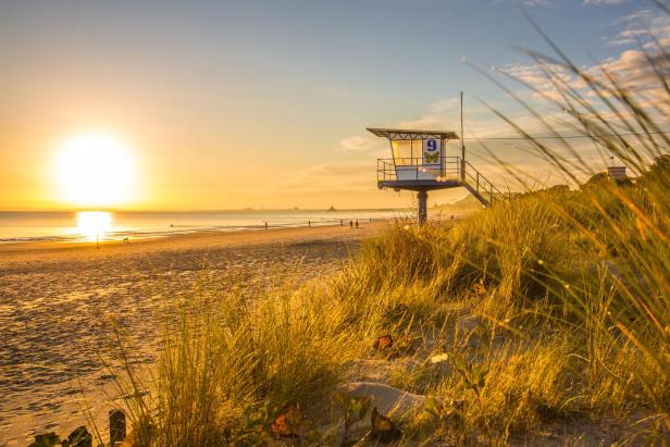 Rettungsturm auf Stelzen am Strand bei Sonnenuntergang, umgeben von Dünengras und mit wenigen Personen am Wasser.
