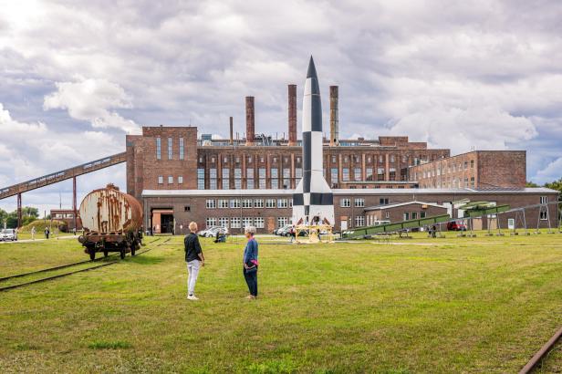 Zwei Personen stehen auf einer Wiese vor einem großen, rotbraunen Backsteingebäude mit mehreren Schornsteinen und einer schwarz-weißen Rakete in der Mitte.