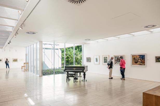 Heller Ausstellungsraum mit Flügelklavier in der Mitte, großen Fenstern und mehreren gerahmten Bildern an den Wänden, zwei Personen betrachten Kunstwerke.