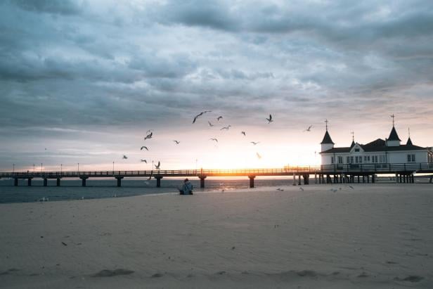 Strand mit Sand, im Hintergrund eine Seebrücke mit Gebäuden, darüber bewölkter Himmel und fliegende Vögel bei Sonnenuntergang.