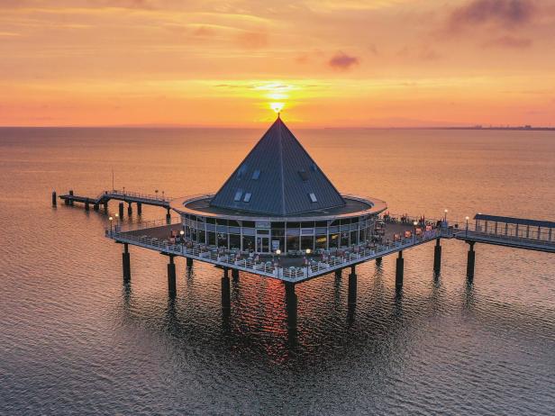 Pyramidenförmiges Gebäude auf Pfählen im Wasser bei Sonnenuntergang mit Steg und Menschen auf der Plattform.