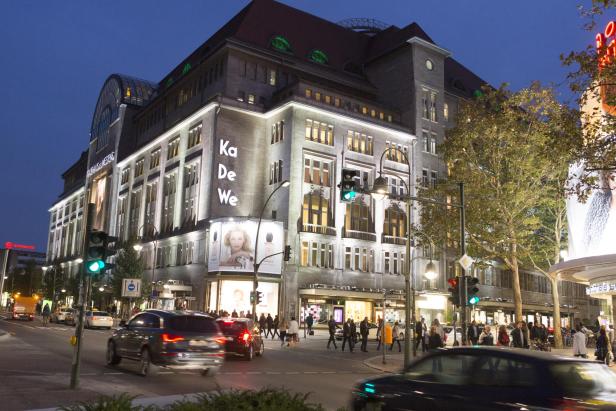 Beleuchtetes Kaufhaus KaDeWe an einer belebten Kreuzung bei Abenddämmerung mit Fußgängern und Autos.