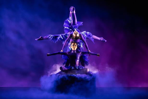 Zwei Tänzer in blauen Kostümen auf einer Bühne mit Nebel, einer sitzt mit ausgestreckten Armen, der andere balanciert kopfüber auf ihm, umgeben von blau-violettem Licht.