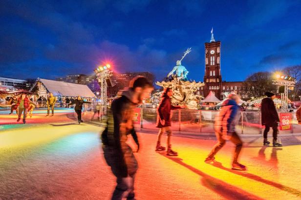 Menschen laufen auf einer beleuchteten Eisbahn vor einem beleuchteten Brunnen und einem Turm bei Abenddämmerung.