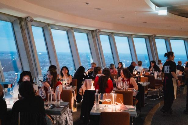 Restaurant mit großen Fenstern und Blick auf die Stadt, Gäste sitzen an gedeckten Tischen, Kellnerin in schwarzer Kleidung bedient Gäste.