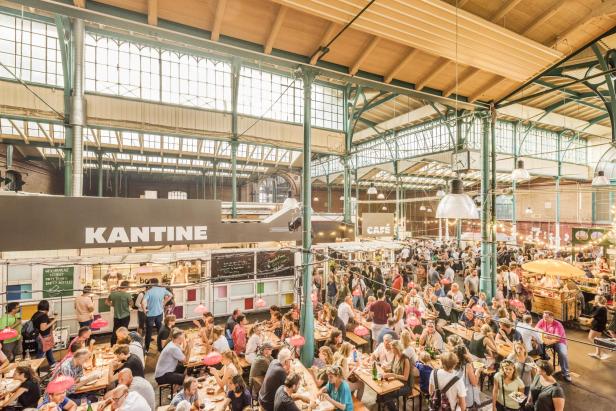 Große, helle Markthalle mit vielen Menschen an Holztischen, Ständen mit den Schildern »KANTINE« und »CAFE« und grünen Stahlträgern unter Glasdach.