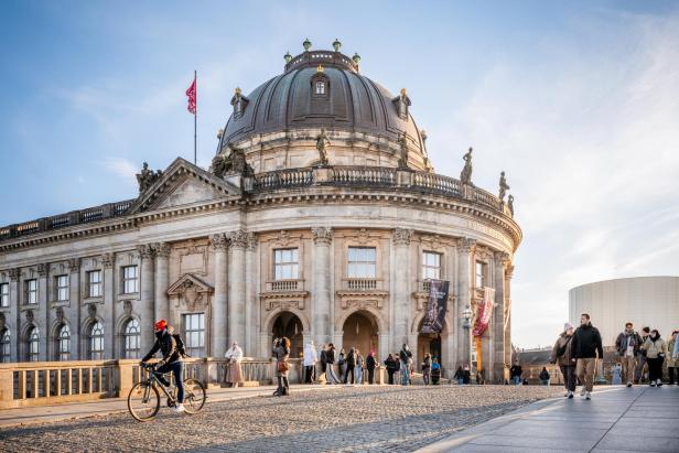 Historisches Rundgebäude mit Kuppel und Flagge, davor Menschen und ein Radfahrer auf gepflastertem Platz bei Tageslicht.