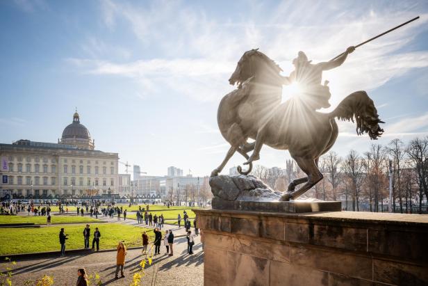 Reiterstatue mit Sonnenschein im Hintergrund, davor Park mit Menschen und historisches Gebäude mit Kuppel.