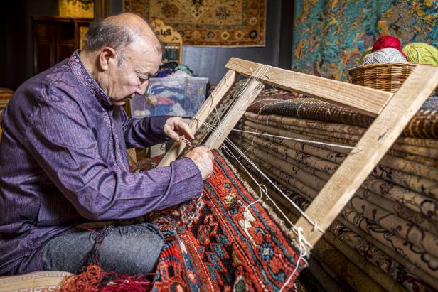 Der Teppichmeister sitzt bei einem Rahmen, darin ist ein löchriger Teppich eingespannt. Er flickt ihn mit einer Nadel und einem Kamm.. Der Mann hockt am Boden, um ihn herum liegen Teppiche aufeinandergestapelt.
