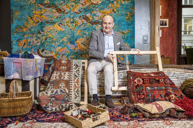 Ali Rahimi sitzt auf einem Berg Teppichen, er hält einen Webrahmen in der Hand, neben ihm liegen Körbe mit Flickzeug. Der Händler trägt Sakko.