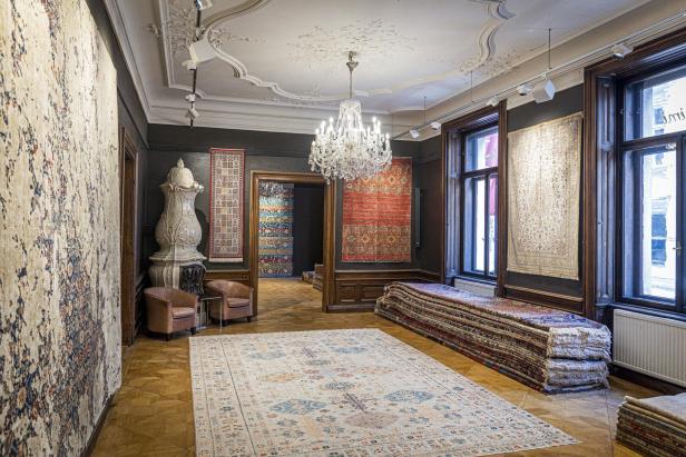 Blick in einen Ausstellungsraum: An den Wänden hängen Teppiche, es gibt Luster, Stuck,  und Teppich liegen schön übereinandergestapelt neben der Wand. 