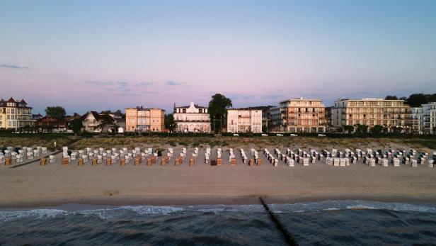 Strand mit Reihen von weißen Strandkörben vor mehrstöckigen Gebäuden unter blauem Himmel.