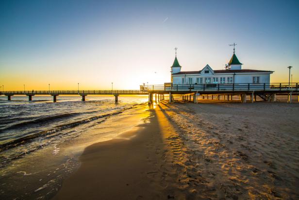 Sonnenuntergang über einem langen Steg mit einem weißen Gebäude mit grünen Türmchen am Ende, Schatten auf dem Sandstrand.