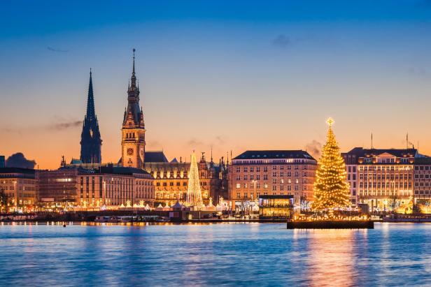 Weihnachtsbaum mit Lichterketten am Ufer eines Flusses vor beleuchteten historischen Gebäuden und Kirchtürmen bei Abenddämmerung in Hamburg