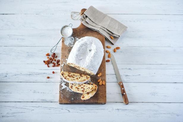 Dresdner Christstollen: Gebackenes Stollenbrot mit Puderzucker auf Holzbrett, daneben Messer, Mandeln, Rosinen, Mehlsieb und Leinentuch auf hellem Holztisch.