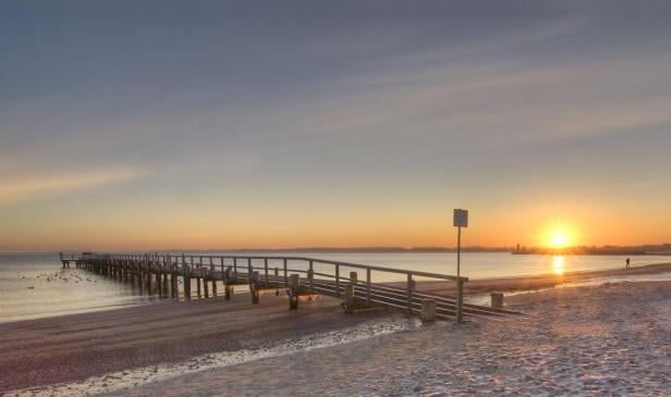 Langer Steg mit Geländer führt über den Strand ins Meer bei Sonnenuntergang mit orangefarbenem Himmel.