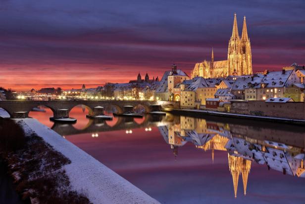 Regensburg: Regensburger Dom und Steinerne Brücke bei Sonnenuntergang mit Spiegelung im Wasser und schneebedeckten Dächern.