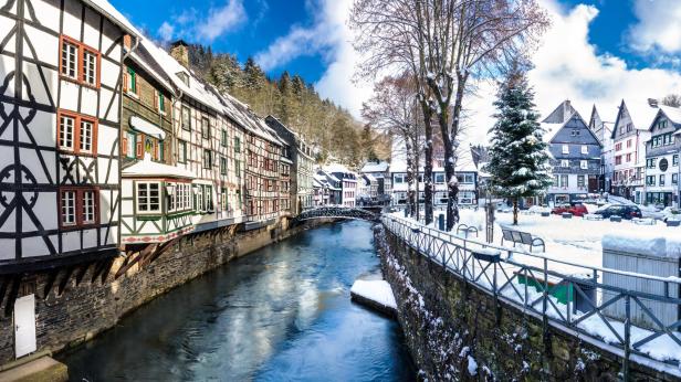 Monschau: Fluss mit Fachwerkhäusern links und schneebedecktem Platz mit Bäumen und Autos rechts unter blauem Himmel