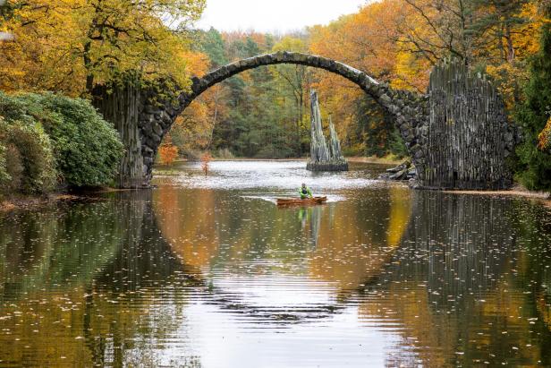 Rakotzbrücke: Steinerne Bogenbrücke über ruhigen Fluss, umgeben von herbstlich gefärbtem Wald, mit Person im Kanu auf dem Wasser.