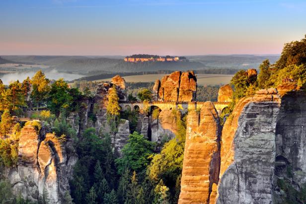 Sächsische Schweiz: Felsformationen mit Bäumen und einer Steinbrücke in einer bewaldeten Landschaft bei Sonnenaufgang.