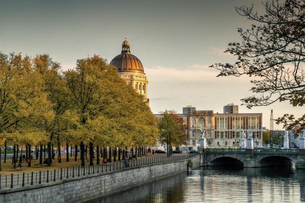 Berlin: Flussufer mit Bäumen in Herbstlaub, dahinter Kuppel eines historischen Gebäudes und Brücke über den Fluss bei klarem Himmel.