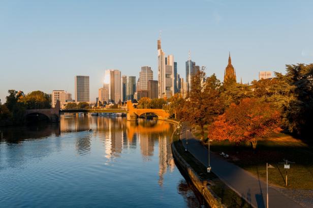 Skyline von Frankfurt am Main mit Hochhäusern und Mainufer im Herbst, Bäume mit herbstlichen Farben, ruhiger Fluss mit Spiegelung.