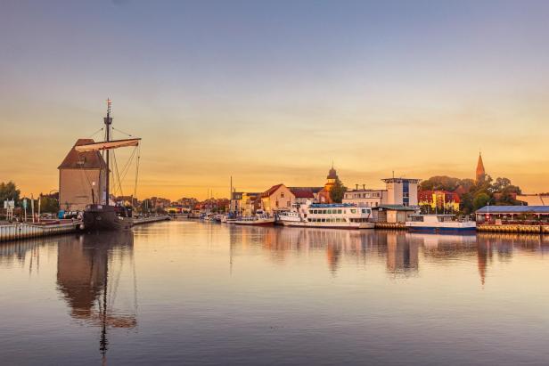 Uckermünde: Hafen mit altem Segelschiff und Passagierboot bei Sonnenuntergang, Gebäude und Kirchturm im Hintergrund.