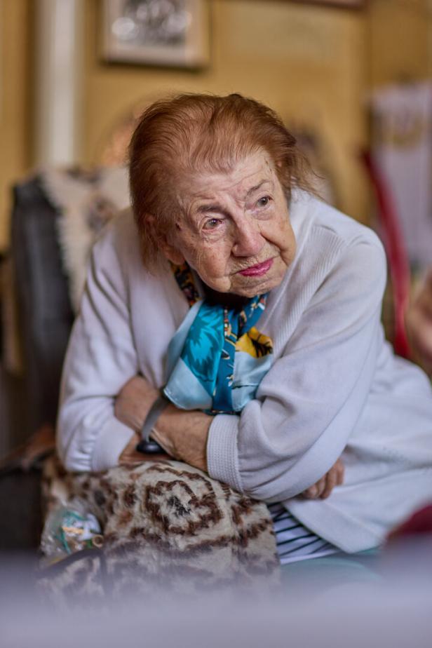 Influencerin Erni Jasek, 100 Jahre alt, sitzt mit heller Strickjacke und buntem Halstuch sitzt nachdenklich mit verschränkten Armen auf einem Sofa.