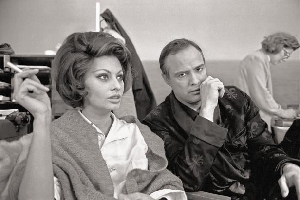 Am Set mit Marlon Brando. Beide Sitzen im Stuhl, sie raucht, er sieht ernst in die Kamera.