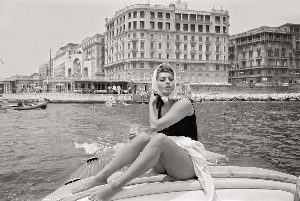 Sophia Loren sitzt mit Badeanzug und Kopftuch auf einem Boot.