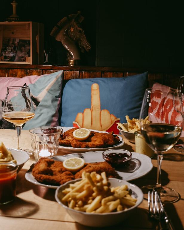 Ein gedeckter Tisch mit Schnitzel, Pommes, Wein. Im Hintergrund ist ein Polster zu sehen, auf dem ein Stinkefinger prangt.