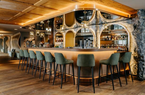 Eine stilvolle Bar mit grünen Barhockern, beleuchtetem Tresen und dekorativer Stein- und Spiegelfront.