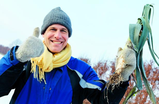 Person in blauer Jacke, grauer Mütze und gelbem Schal hält zwei Lauchknollen, zeigt Daumen hoch.