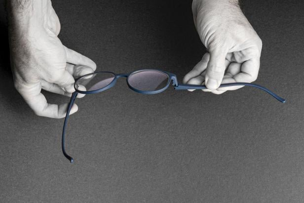 Zwei Hände halten eine Brille mit runden Gläsern und dünnem, dunklem Gestell vor dunklem Hintergrund.