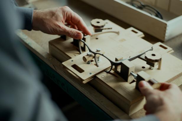 Hände halten eine schwarze Brille über eine hölzerne Werkbank mit mechanischen Bauteilen.
