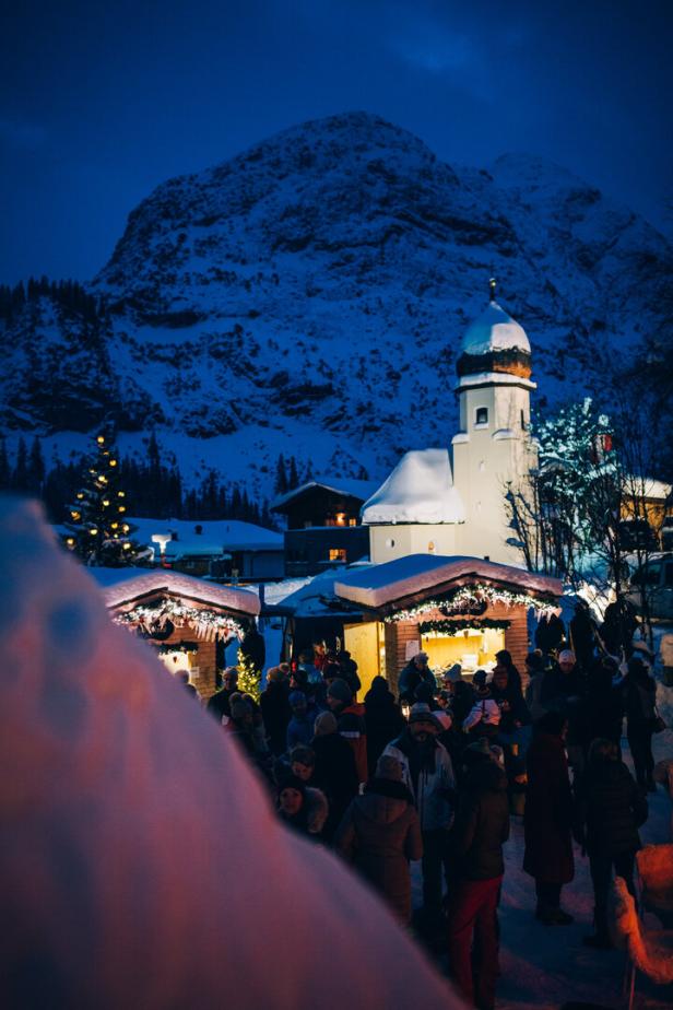 Weihnachtsmarkt mit beleuchteten Ständen und Menschen vor schneebedeckter Kirche und Bergkulisse bei Abenddämmerung.