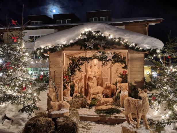 Krippe aus Strohfiguren mit Maria, Josef, Jesus und mehreren Tieren in einer beleuchteten Holzhütte im Schnee bei Nacht.
