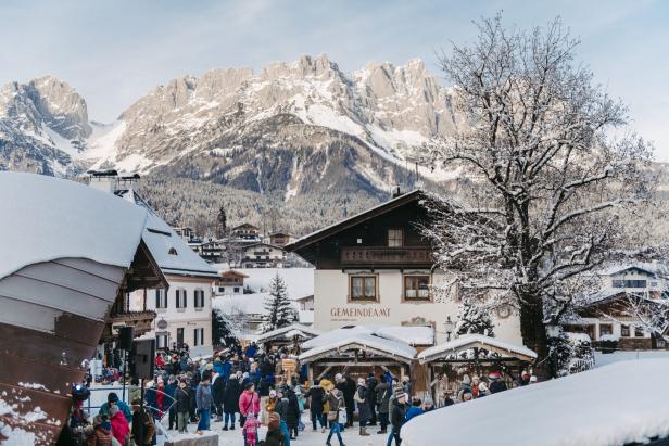 Winterliche Berglandschaft mit schneebedeckten Häusern und vielen Menschen vor einem Gebäude mit der Aufschrift 'GEMEINDEAMT'.